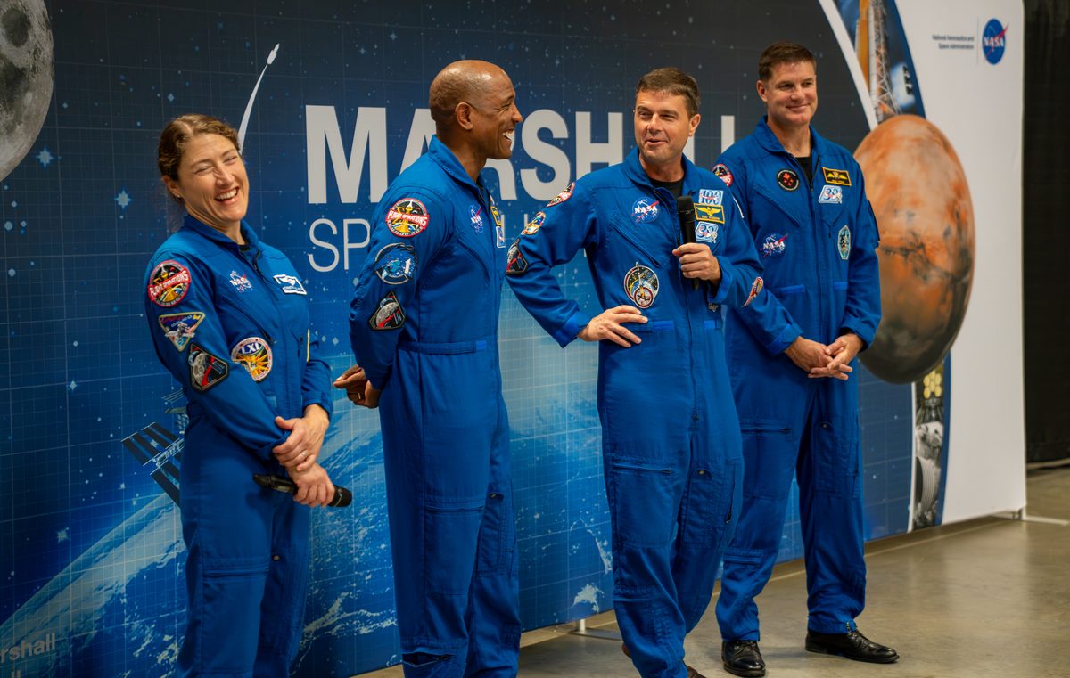 The Artemis II crew smile and laugh. They are wearing blue jumpsuits with patches on them. Christina Koch, left, and Reid Wiseman, second from right, are holding microphones.