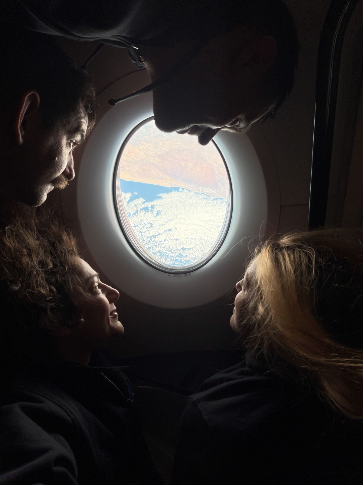 NASA’s SpaceX Crew-12 crew members surround an oval-shaped window aboard a SpaceX Dragon, looking out at clouds above blue water and a tan landmass. From the left, rotating clockwise around the image are NASA astronaut Jack Hathaway, Roscosmos cosmonaut Andrey Fedyaev, ESA (European Space Agency) as