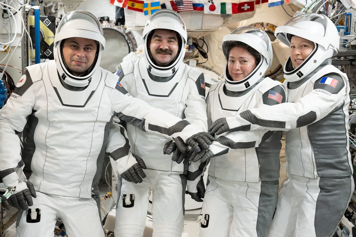 Four astronauts in white space suits smile at the camera and put their gloved hands together aboard the space station.