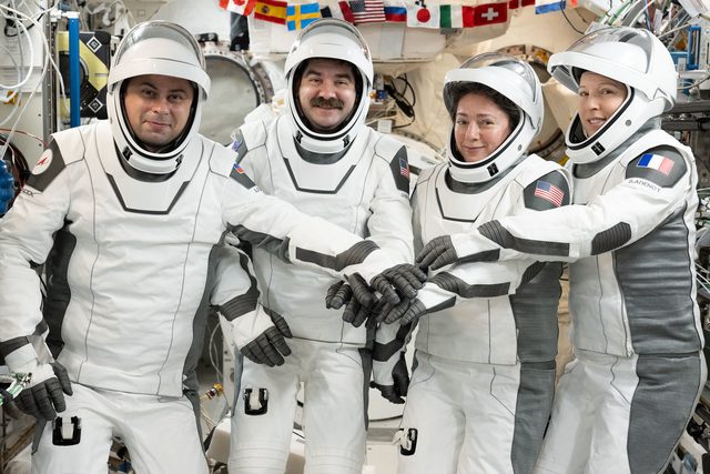 Four astronauts in white space suits smile at the camera and put their gloved hands together aboard the space station.