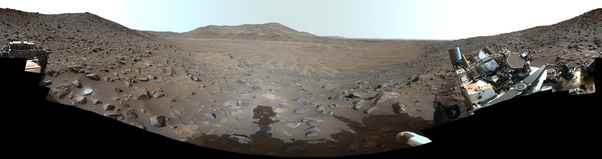 A panoramic view of a barren, rocky landscape with distant hills, framed by parts of a robotic rover visible on the left and right edges.