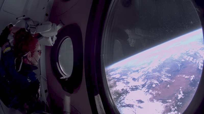 The curve of the Earth and a thin blue band of atmosphere seen through a round cabin window, with a passenger's shoulder in the foreground.