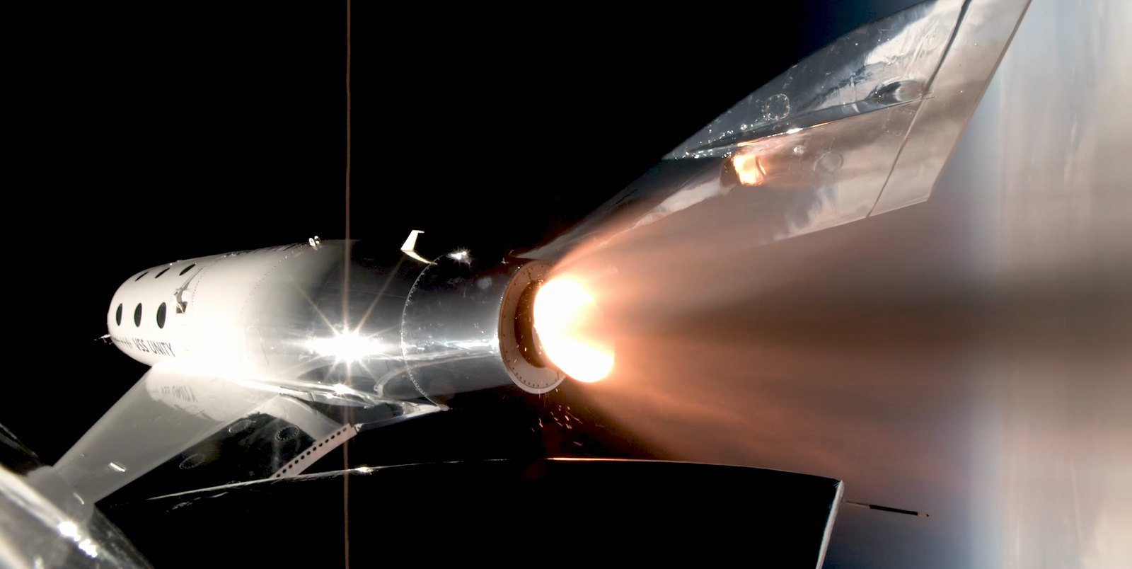 A silver spaceship against the black of space, its rocket motor firing a bright orange plume, seen from a camera on the ship itself.