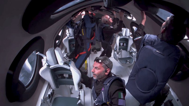 Passengers in flight suits float above their seats in a white cabin lined with round windows.