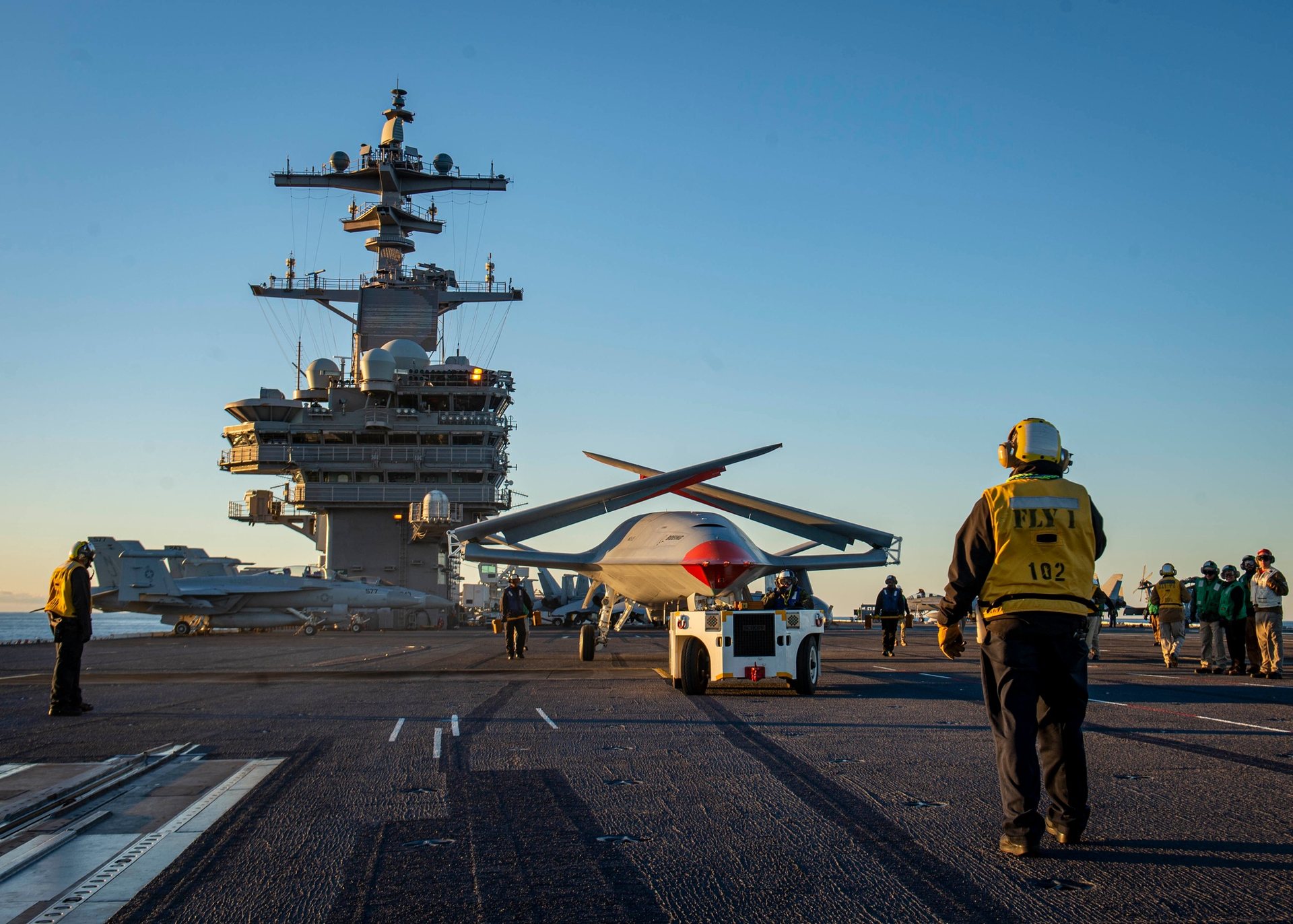 A Boeing MQ-25 test aircraft, wings folded, being directed across the flight deck of the aircraft carrier USS George H.W. Bush at sunrise on December 13, 2021, with the ship's island and Super Hornets behind it. U.S. Navy photograph.